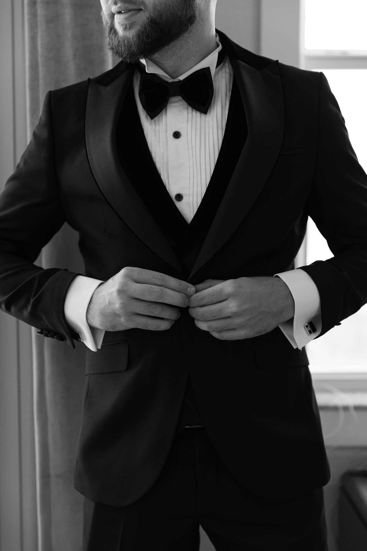 Man adjusting his black tuxedo jacket, wearing a white dress shirt and bow tie, showcasing Bold Italia Suit's luxury customized style experience.