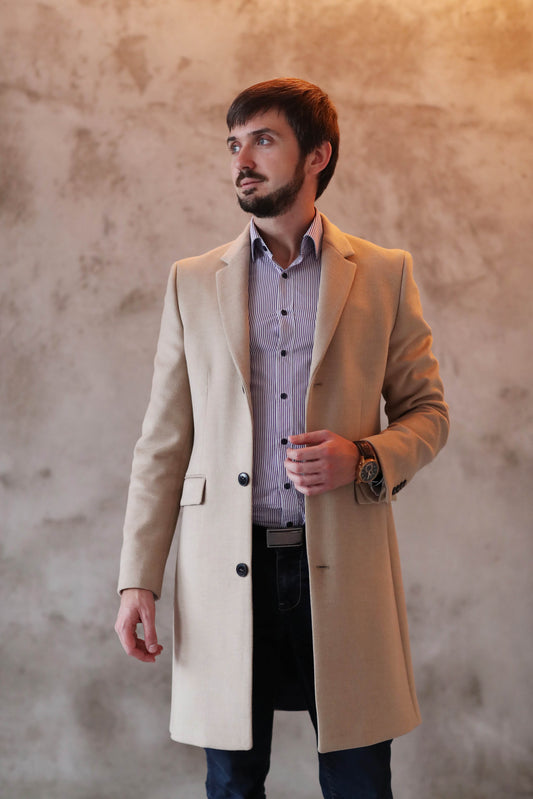 Man wearing a beige custom overcoat from Bold Italia Suit, standing against a textured wall, showcasing a stylish and tailored outerwear option.