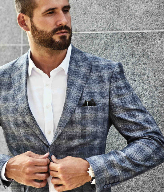 Stylish man wearing a modern gray plaid suit with a white dress shirt, adjusting his jacket against a sleek stone wall backdrop, showcasing premium menswear from Bold Italia Suit.