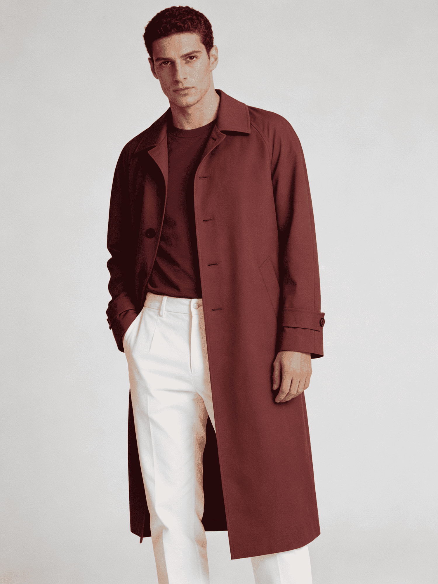 Man wearing a brown trench coat over a dark shirt and white pants on a light gray background