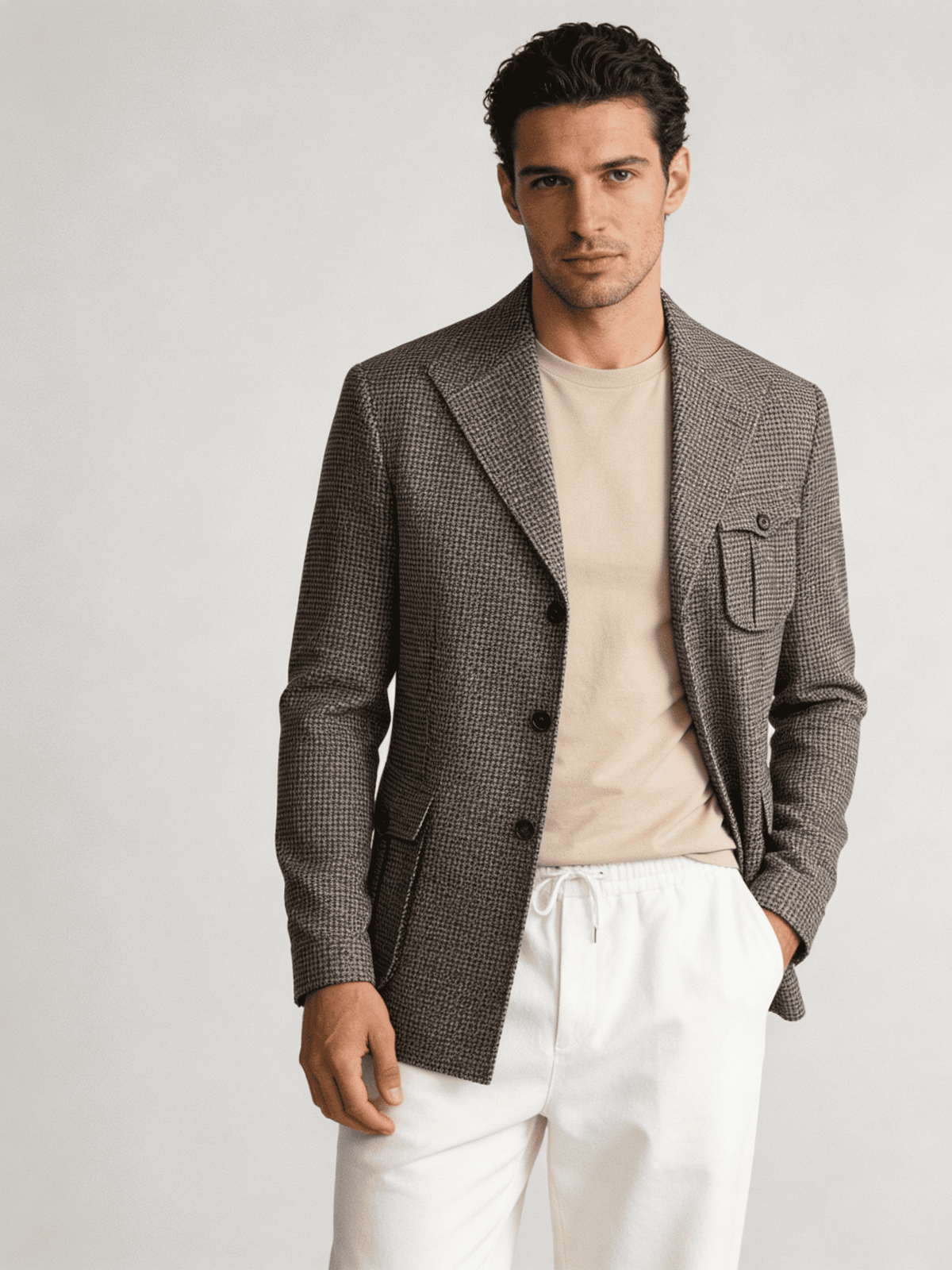 Man wearing a brown blazer over a beige shirt with white pants on a light gray background