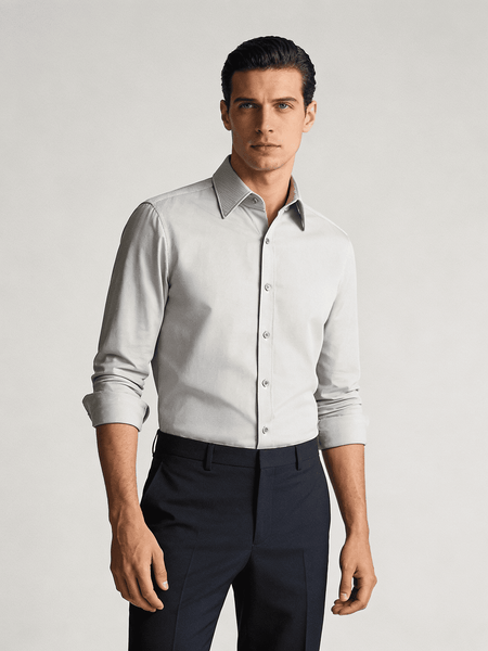 Man wearing a light gray dress shirt with black trim and dark pants on a plain background