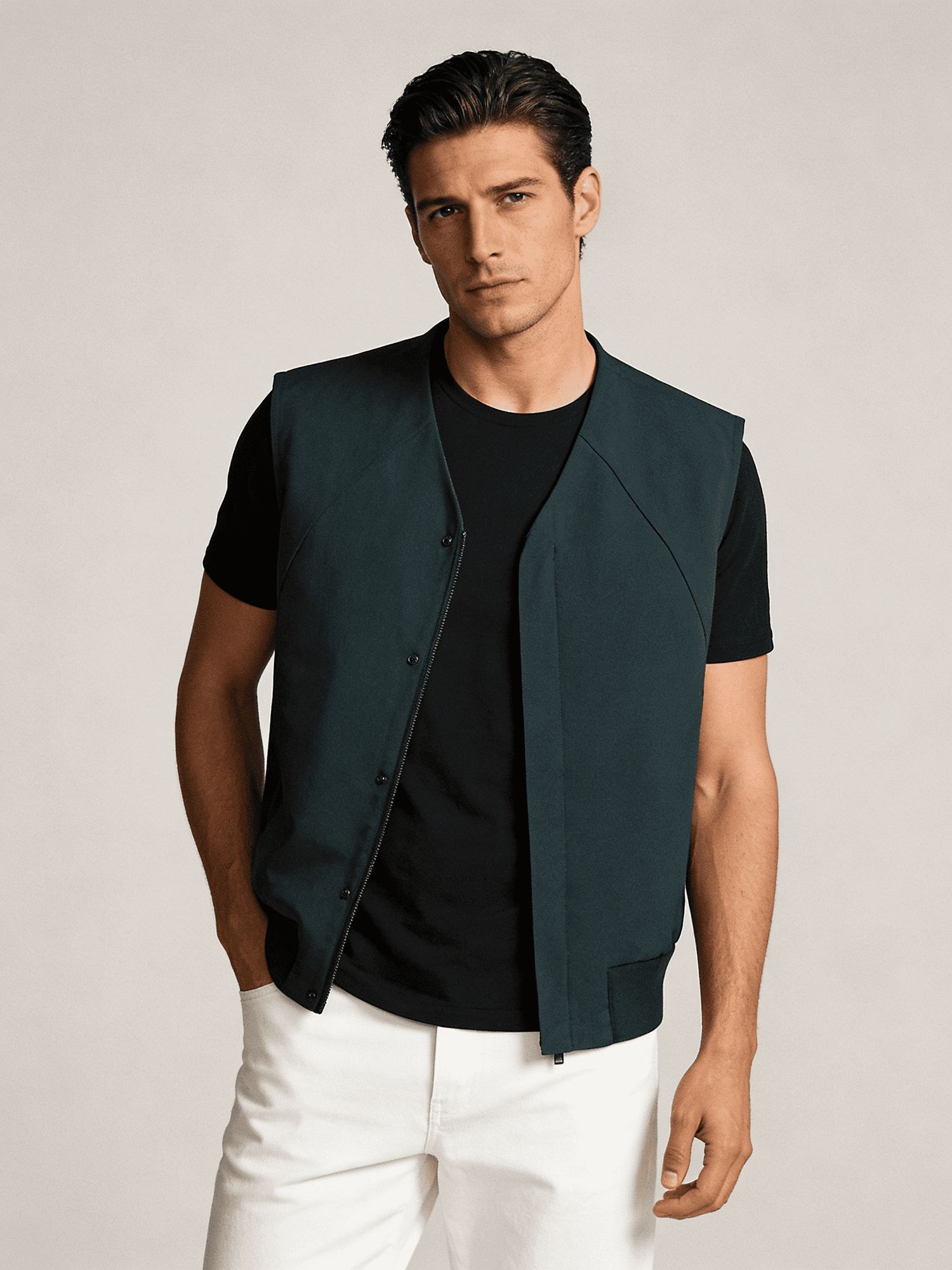 Man wearing a dark green vest over a black shirt with white pants on a light gray background